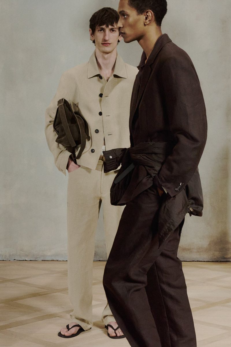 Two fashion models stand indoors on a light-colored floor and wear dark and light brown toned, casual jackets and pants with flip-flops.