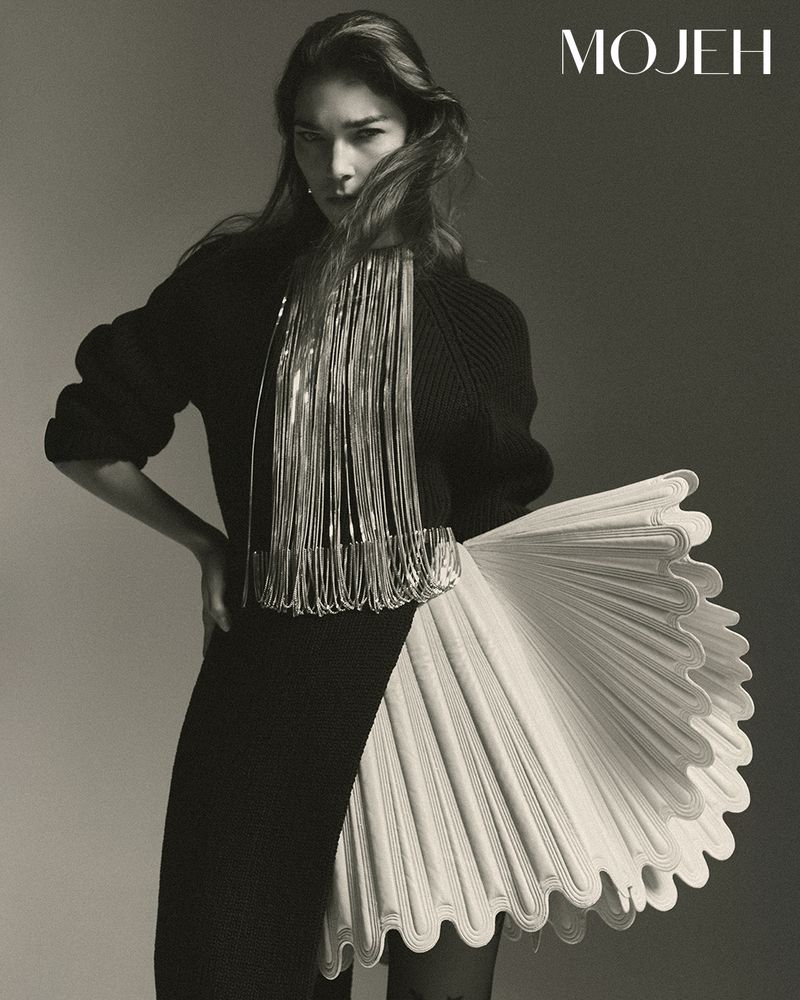 Fashion editorial shot of an adult woman in a black knit dress with decorative fringe and ruffled skirt element, posing in a studio.