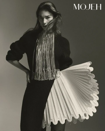 Fashion editorial shot of an adult woman in a black knit dress with decorative fringe and ruffled skirt element, posing in a studio.