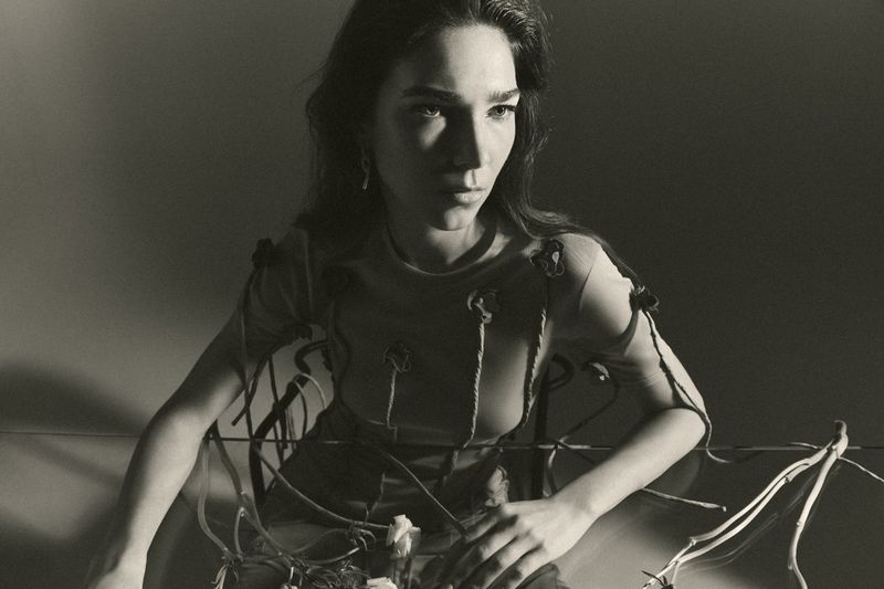 Monochrome portrait of a young woman with long hair, wearing a flower-embellished top, seated in front of a reflective surface.