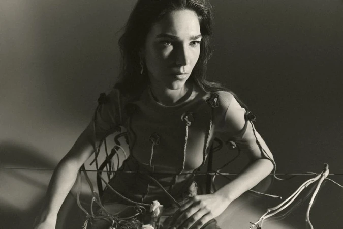 Monochrome portrait of a young woman with long hair, wearing a flower-embellished top, seated in front of a reflective surface.