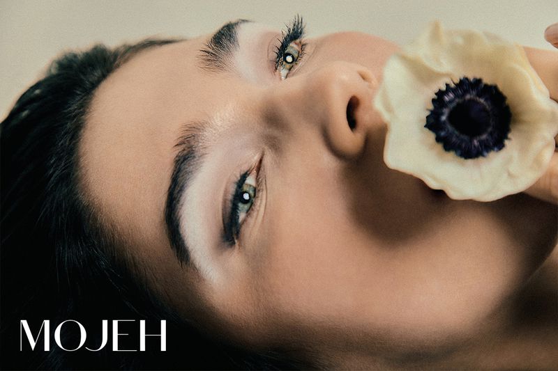 Close-up portrait of an adult woman with light skin and green eyes, with a white flower near her face and the brand name 'MOJEH' visible.