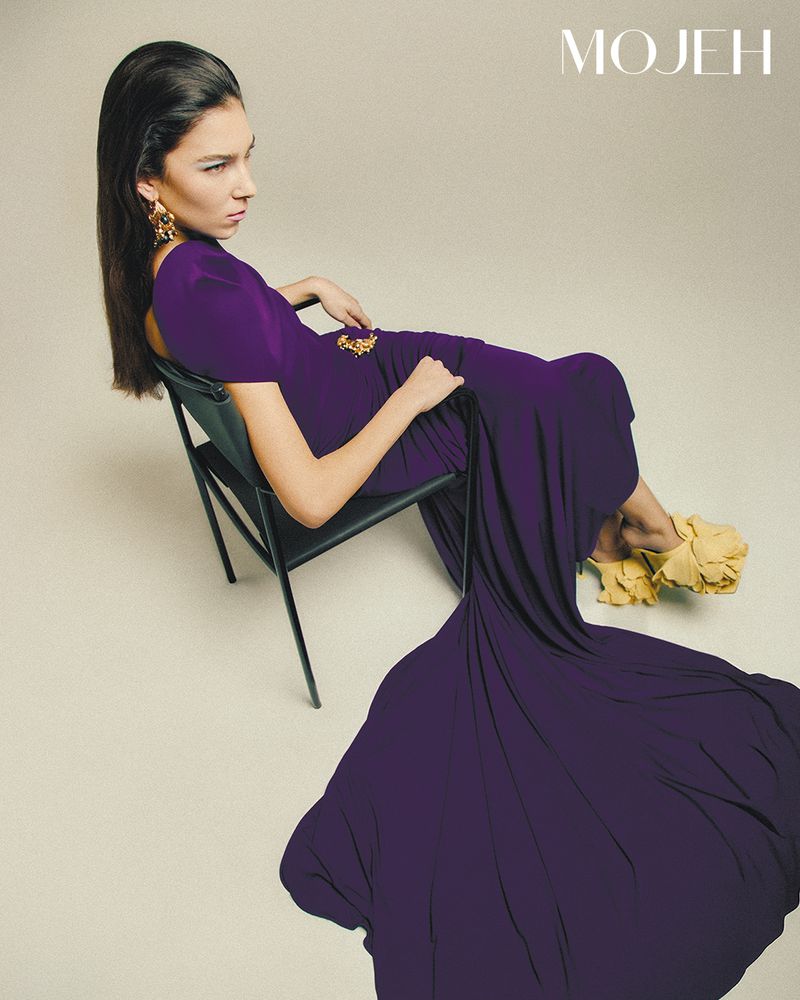 Fashion editorial shot of a female model in a purple gown and yellow shoes reclining on a black folding chair in a light studio.