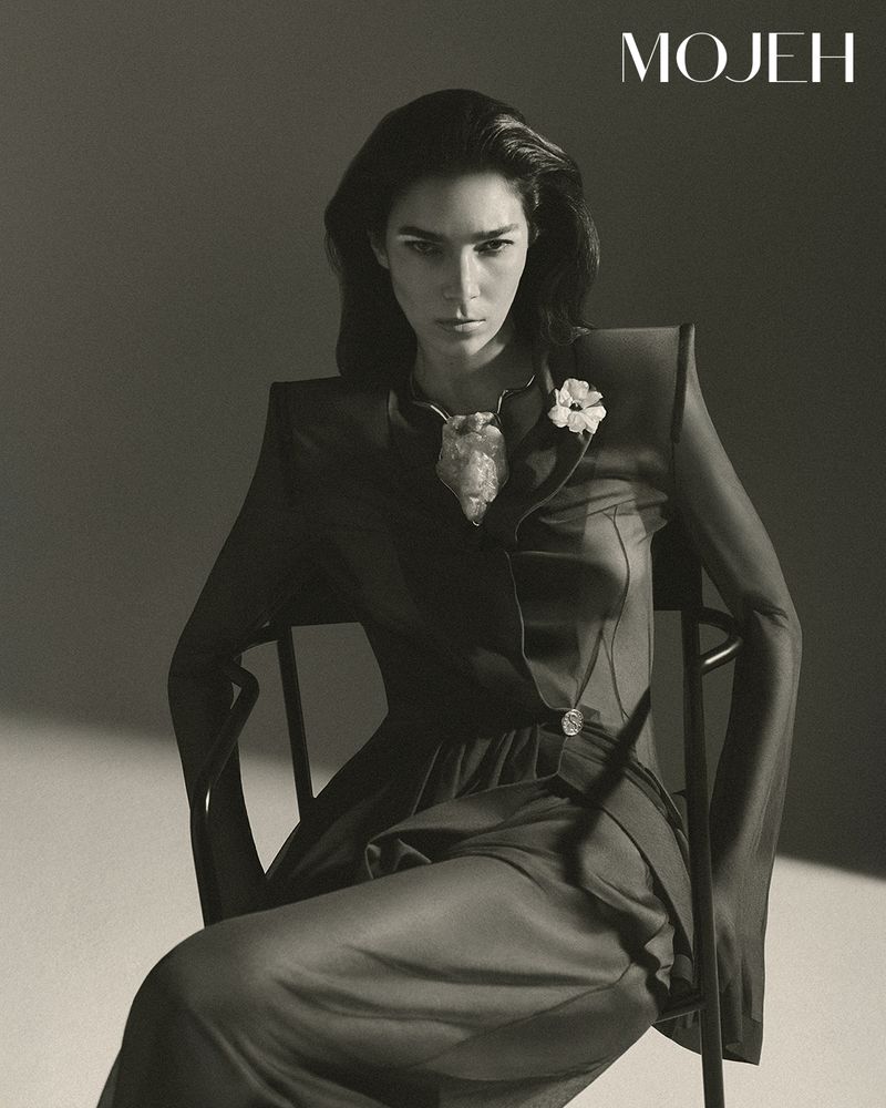 Portrait of a woman with dark hair, wearing a black dress, seated in a chair, jewelry details, set against a neutral background.