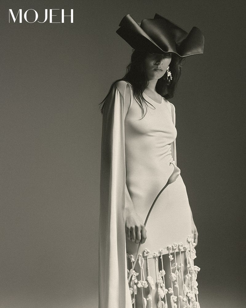Monochrome fashion shot of an adult female model wearing a light-colored dress, oversized hat, and earrings, posing in a photo studio.