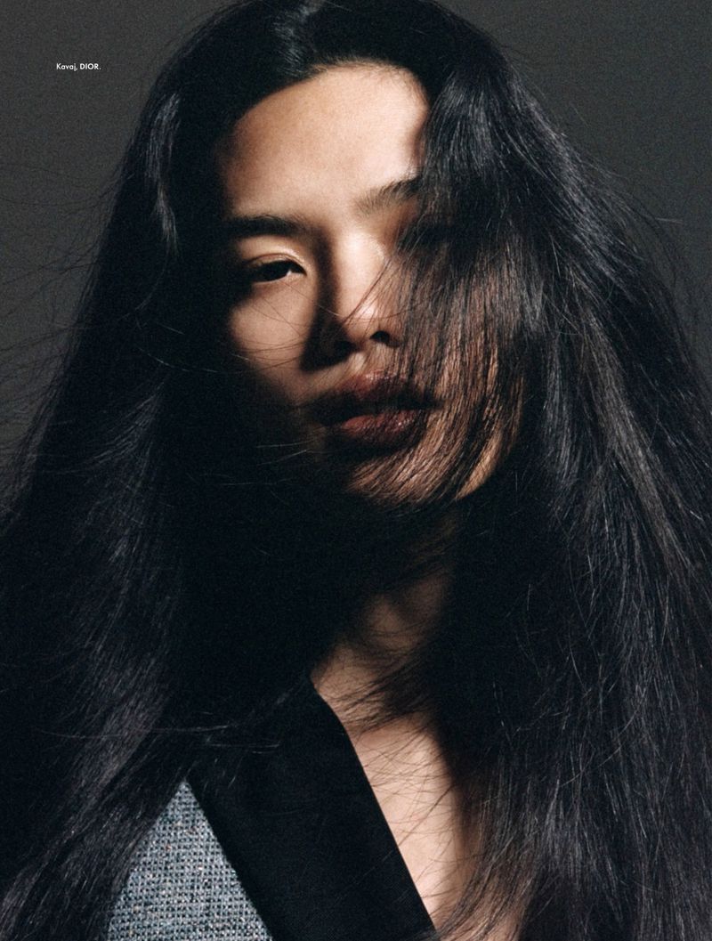 Portrait of a person with long, dark hair wearing a blazer, set against a dark backdrop, photographed in a studio-lit environment.