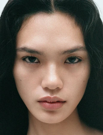 Close-up portrait of a young adult woman with long, dark hair and light skin, gazing directly at the camera with a neutral expression.