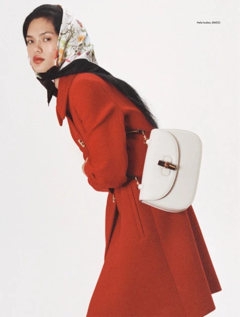 Editorial fashion shot: woman in a red coat and floral headscarf poses in a studio with a white leather purse, clean white backdrop.