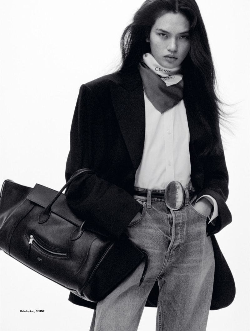 Fashion editorial shot of a long-haired woman in jeans, blazer, and neck scarf holding a black leather bag in a white studio.