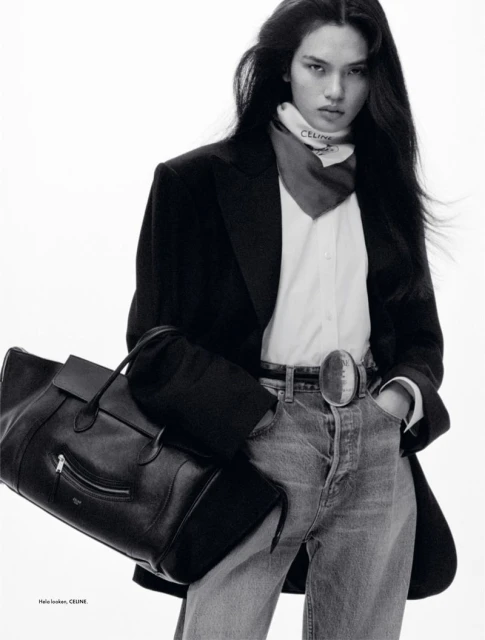 Fashion editorial shot of a long-haired woman in jeans, blazer, and neck scarf holding a black leather bag in a white studio.