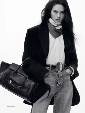 Fashion editorial shot of a long-haired woman in jeans, blazer, and neck scarf holding a black leather bag in a white studio.