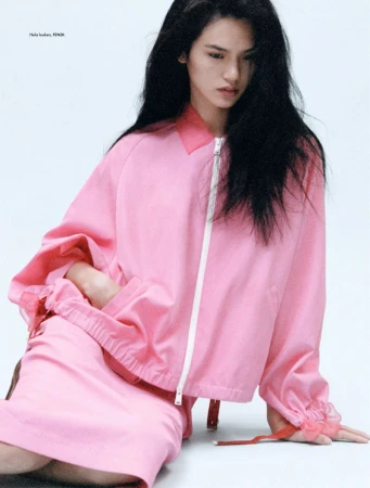 Asian woman in a pink jacket and skirt poses indoors against a white background, showcasing a modern and stylish fashion aesthetic.