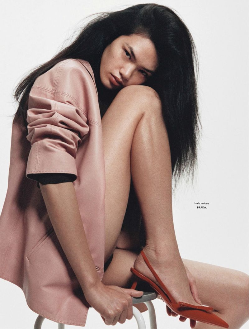 Brunette poses on a stool wearing a pink blazer and red slingbacks in a clean studio fashion shot with strong lighting and bold colors.