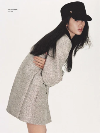 Elegant woman posing in a studio wearing a gray coat dress and black cap. The model has long, dark hair and is looking at the camera.