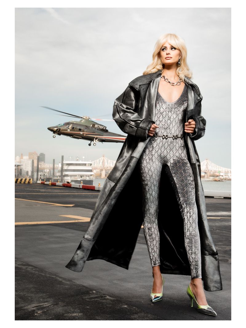 A blonde woman in a patterned catsuit and coat poses on a tarmac with a helicopter and city skyline in the background. Metallic sheen.