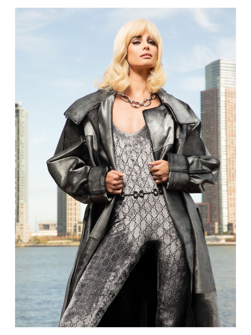Model Taylor Hill wearing a metallic coat and patterned jumpsuit with a city skyline and water background