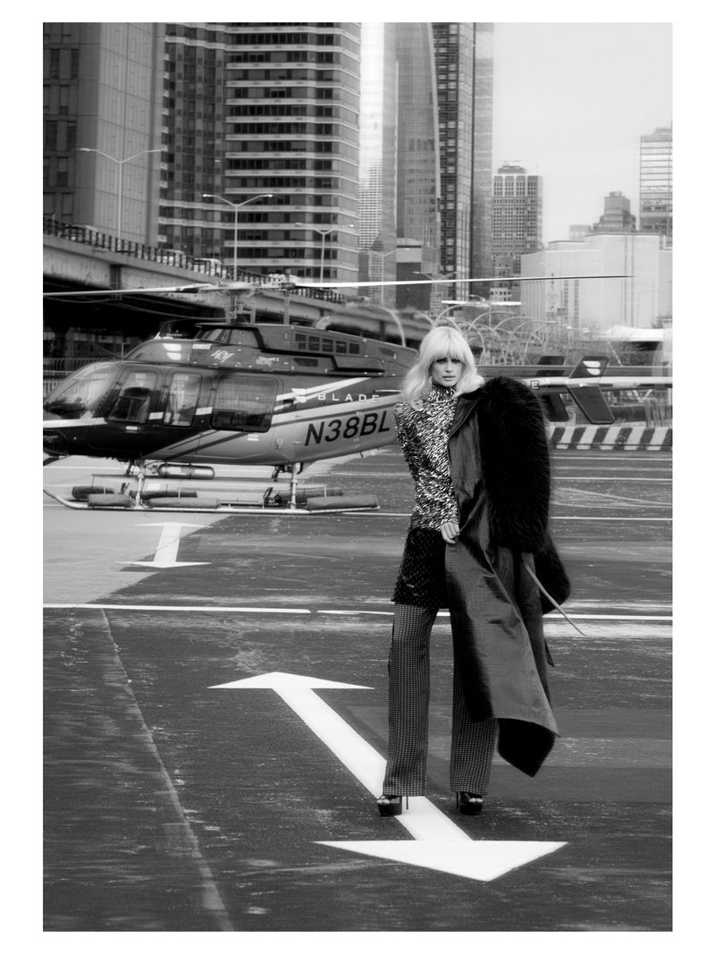 Fashionable woman poses in front of a helicopter on a city helipad, wearing patterned jacket, wide-leg trousers, and holding a coat.