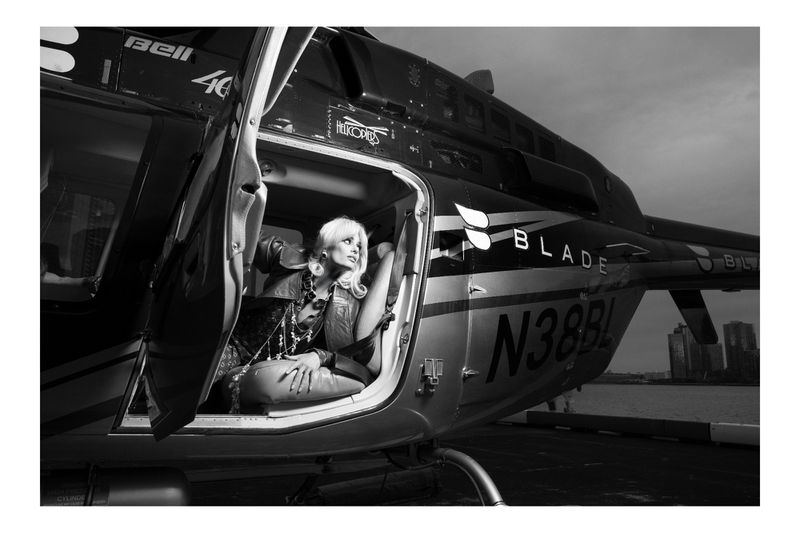 Black and white photograph of a blonde woman seated inside of a helicopter, wearing a jacket and jewelry, gazing out the window.