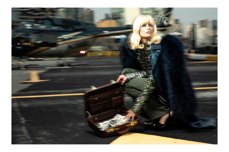 Blonde woman in fur coat squats with briefcase full of cash on airport tarmac with blurred helicopter and city skyline in background.