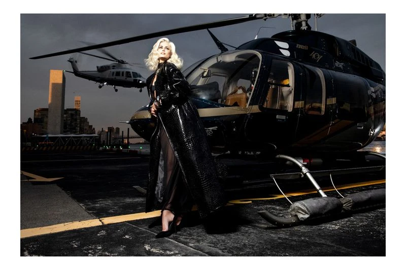 Blonde woman poses in a black, shiny trench coat on a helipad with a helicopter and city skyline in the background at night.