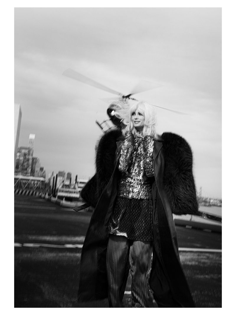 A woman with blonde hair in a shiny top and pants stands outdoors near a helicopter with city buildings in the background in a black and white photo.