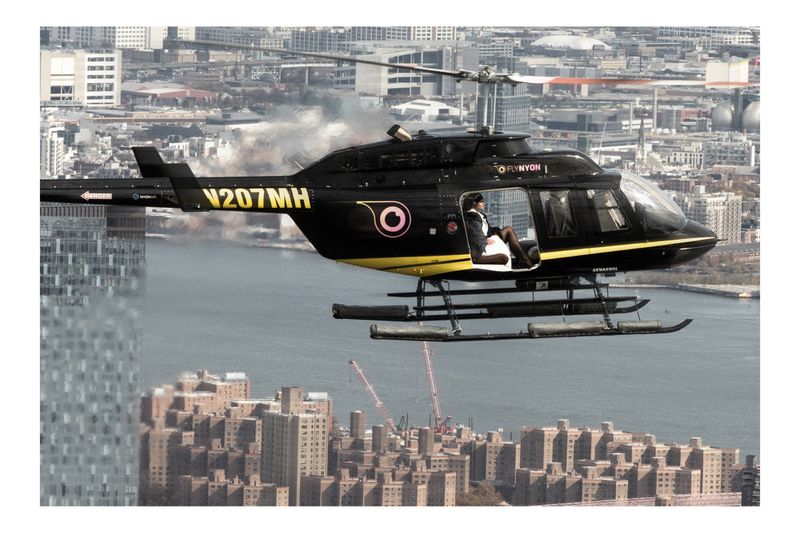 Black and yellow helicopter with passenger flying over a city skyline, featuring modern buildings and a river in the background.