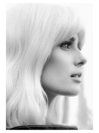 Black and white portrait of a woman with blonde wavy hair, looking off to the side in a photo studio with soft, diffused lighting.