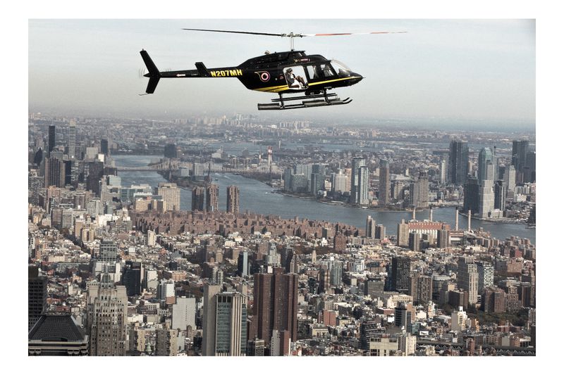 Helicopter flies over a densely populated urban cityscape with numerous tall buildings and waterways on a hazy day.