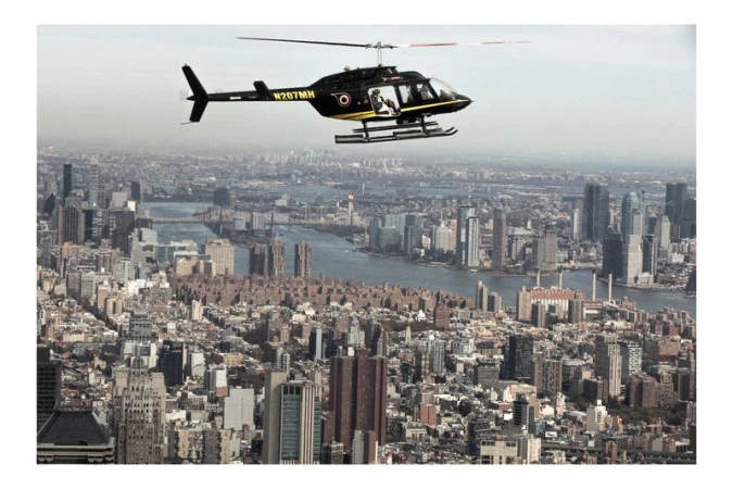Helicopter flies over a densely populated urban cityscape with numerous tall buildings and waterways on a hazy day.