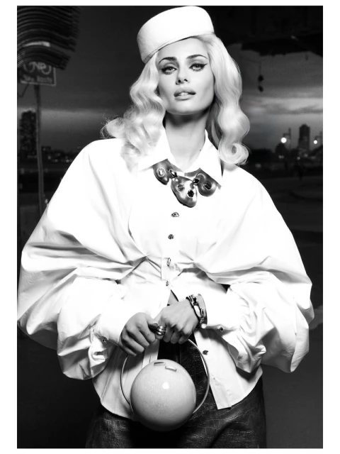 Monochrome portrait of a stylish woman in a white blouse and hat, holding a round bag against an urban skyline backdrop.