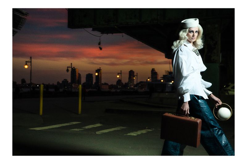 Model Taylor Hill wearing a white sailor hat, white shirt, and dark trousers walking outdoors at dusk