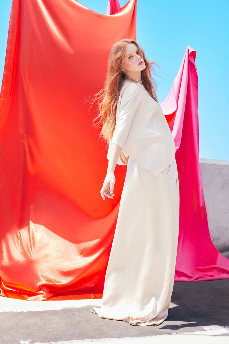 A woman in an off-white dress poses outdoors on a concrete rooftop in front of draped red and pink fabrics against a blue sky.
