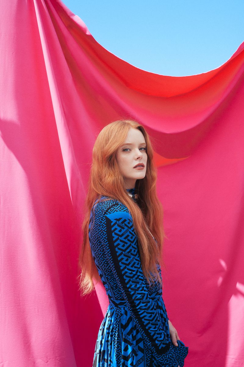 Attractive red-haired woman poses in a blue patterned dress against a vibrant pink curtain and a clear blue sky in an outdoor setting.
