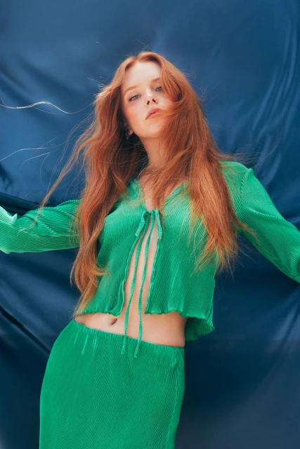 A red-haired woman in a green pleated outfit poses against a blue backdrop, showcasing a modern fashion aesthetic.
