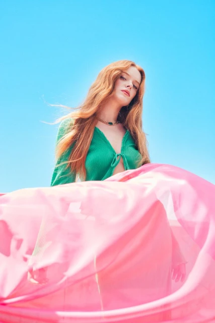 Red-haired woman in green top with pink fabric against a bright blue sky, portrait shot, fashion and beauty concept.