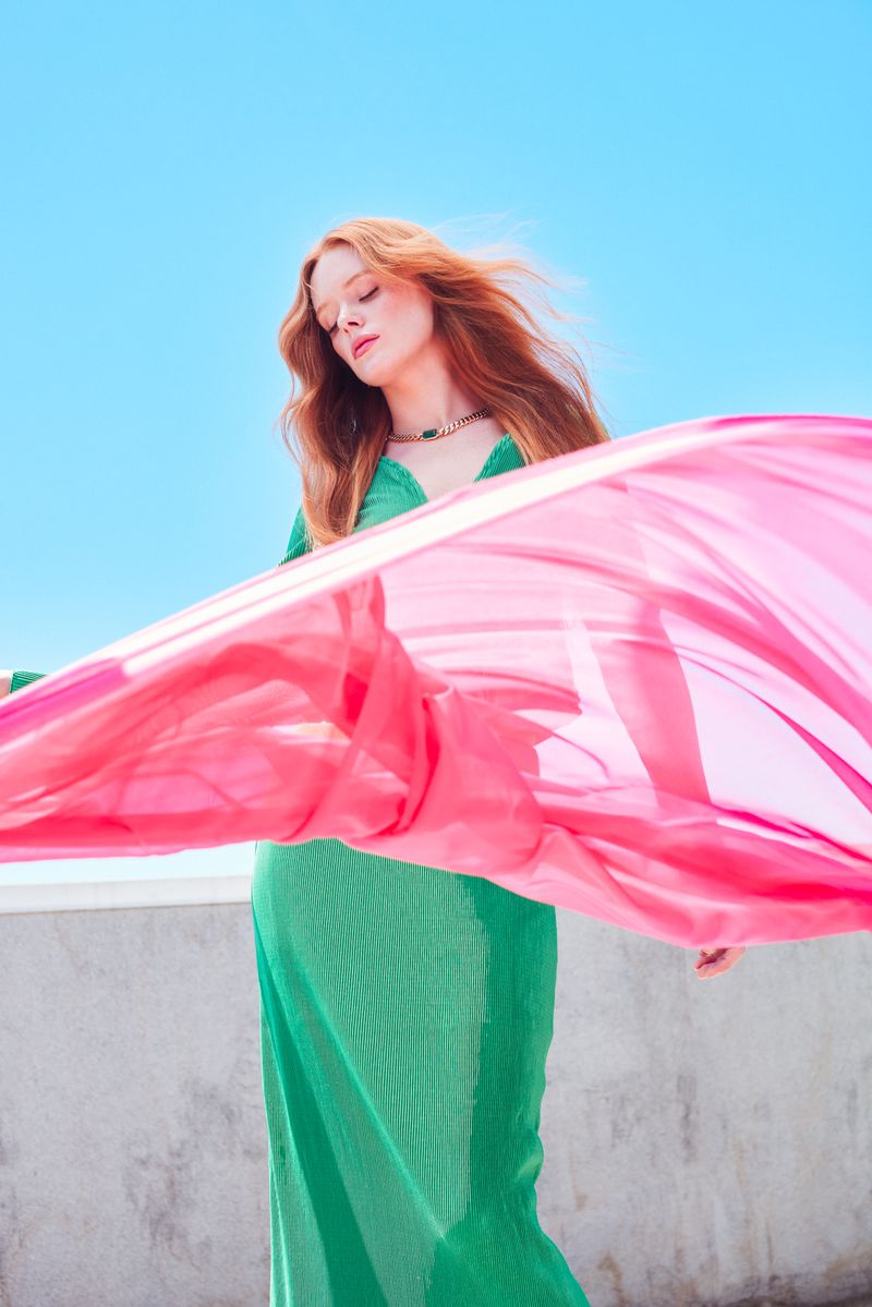 Woman in green dress outdoors with pink fabric blowing in the wind under a blue sky, creating a vibrant and airy fashion portrait.