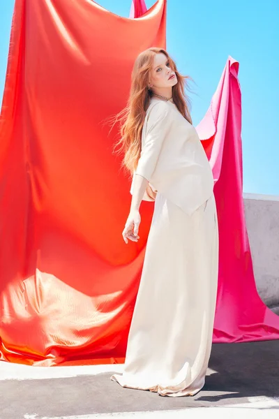 Red-haired woman in cream-colored top and long skirt posing against bright red and pink hanging fabric backdrops.