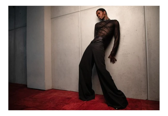 Fashionable black male model poses indoors, wearing a sheer black long-sleeved top and wide-leg black trousers, leaning against a textured grey wall.