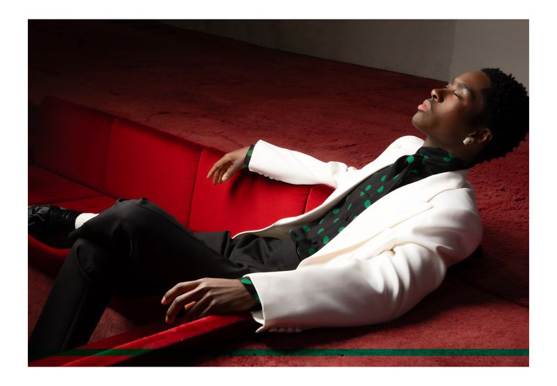Adult Black male model reclines on a red velvet couch wearing a white blazer, black pants, and a black-and-green polka dot shirt in studio.