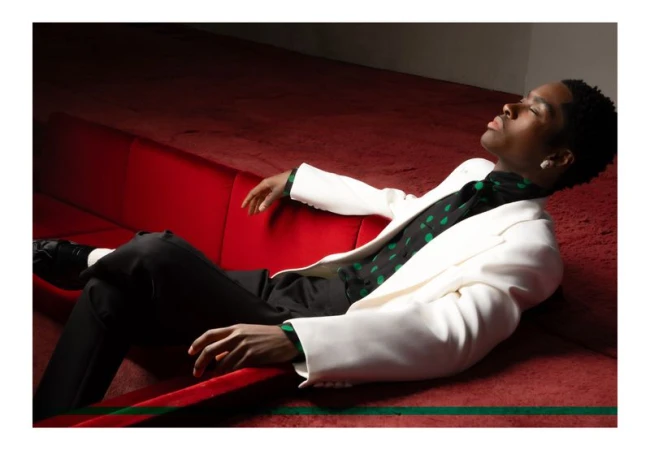 Adult Black male model reclines on a red velvet couch wearing a white blazer, black pants, and a black-and-green polka dot shirt in studio.