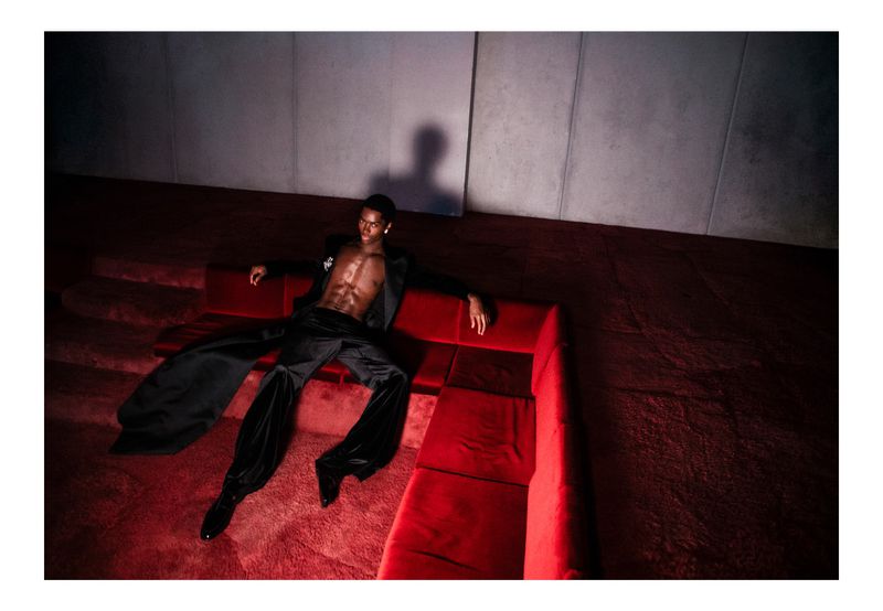 An adult male with a bare torso, wearing black trousers and a blazer, poses on a red modular couch in a dimly lit indoor setting.