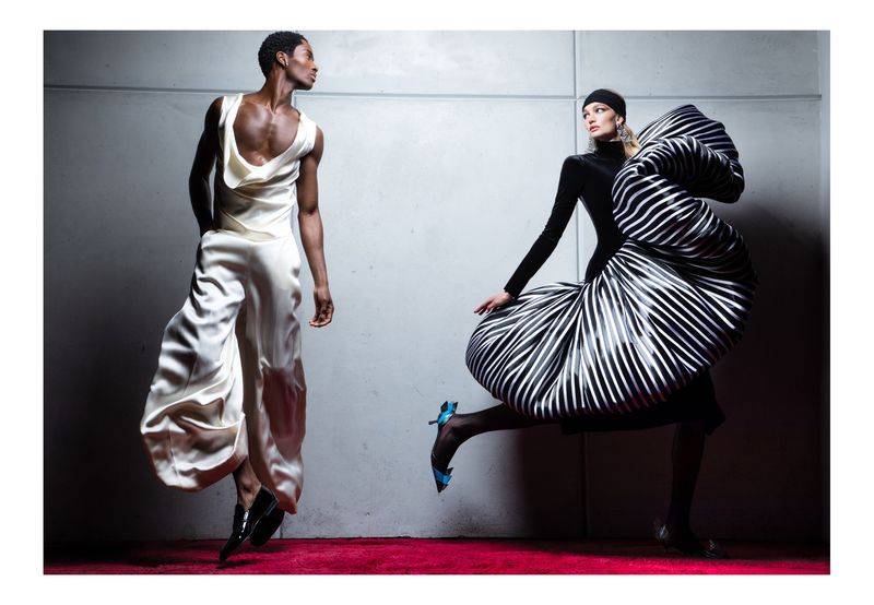 Two fashion models strike dynamic poses in a studio, one in white satin, the other in a black and white striped voluminous gown.