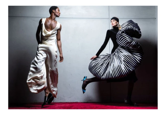 Two fashion models strike dynamic poses in a studio, one in white satin, the other in a black and white striped voluminous gown.