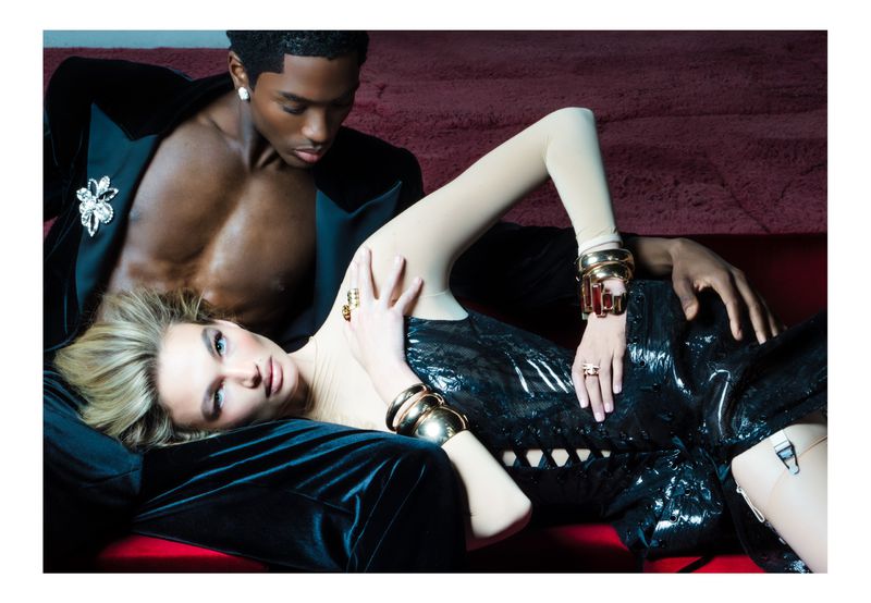 A couple lounges in a stylish photo. An African American man in a suit jacket with an ornamental brooch, and a blond woman with bracelets.