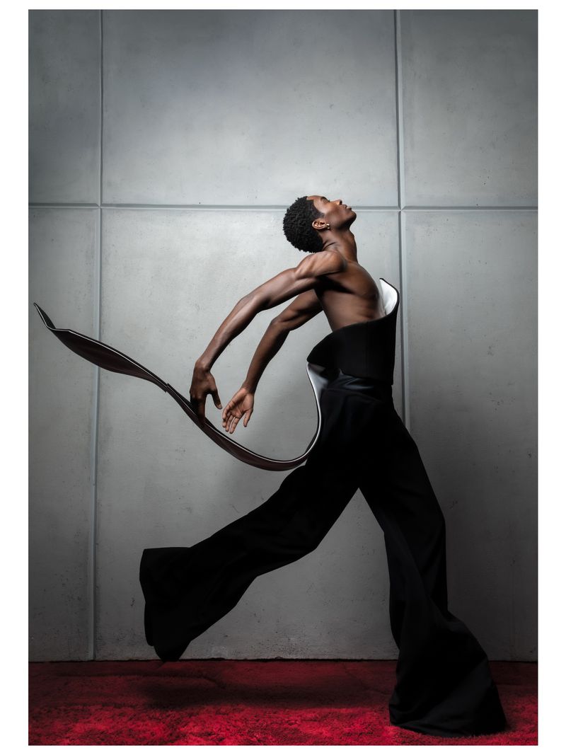 Dramatic studio shot: African-American male dancer in motion with black formal wear, captured in dynamic pose against a gray textured background.