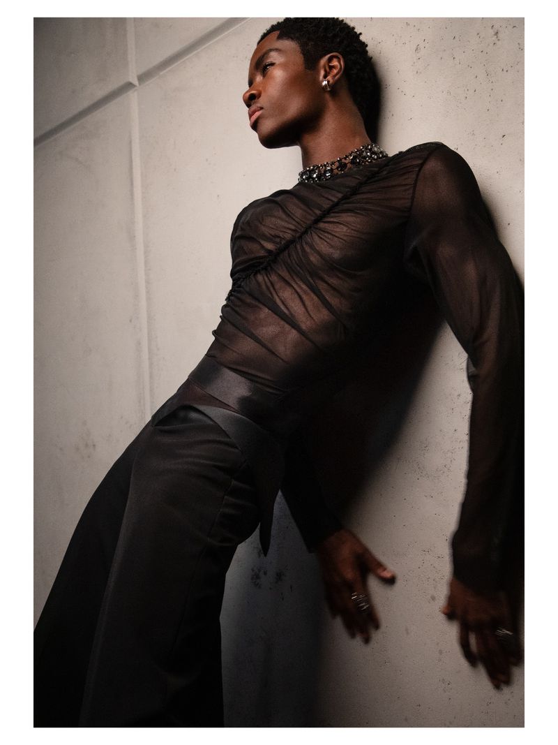Portrait of a Black male model with short hair leaning against a concrete wall, wearing a sheer black top and satin pants in a studio.