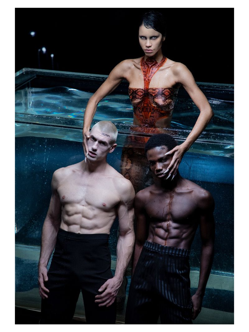 Striking studio shot of a woman standing behind a glass tank with two bare-chested men, set against a stark black backdrop.