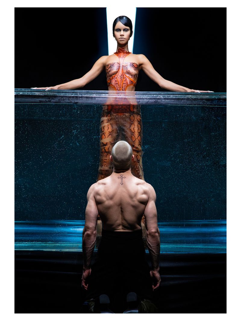 A woman with body art above an aquarium-like container and a shirtless man facing away from the camera below.