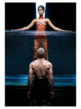 A woman with body art above an aquarium-like container and a shirtless man facing away from the camera below.