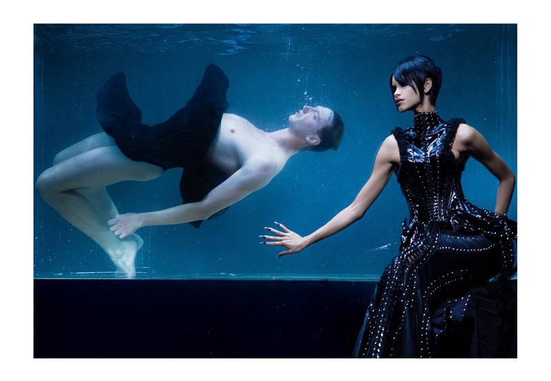 Underwater fashion shot of a woman in a black dress and a man in black fabric, set in clear blue water inside of a glass enclosure.